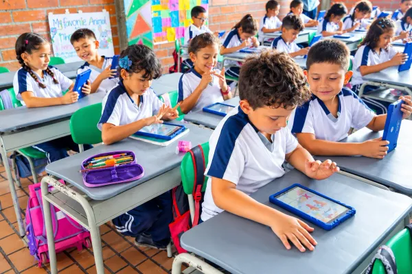 Primary school students in uniform work intently on their tablets, each at their own desk, in a classroom with a brick wall