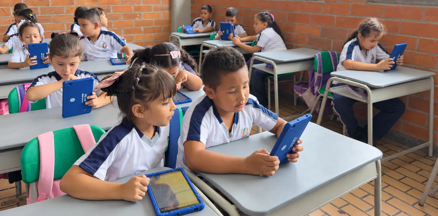 A brick-walled primary classroom with a dozen students at grey desks, each holding a tablet in a blue case