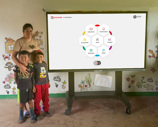 A woman and two young children standing beside an interactive screen showing the AVACOM CLASSROOM menu, in a rural classroom with hand-painted walls