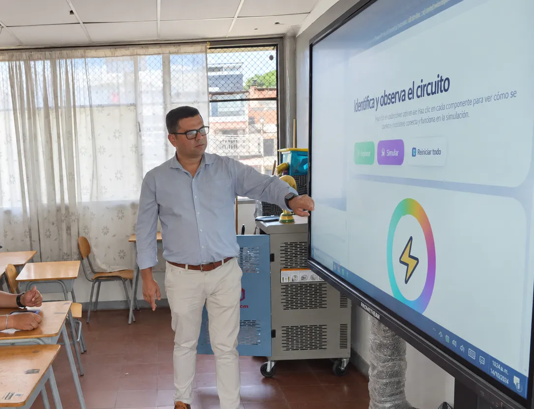 A teacher reaches out to an interactive screen on a wheeled stand showing an activity titled 'Identifica y observa el circuito'