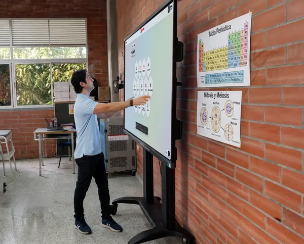 Teacher in a light blue polo shirt reaching out to touch an interactive screen showing the AVACOM CLASSROOM menu, in an empty classroom with a brick wall