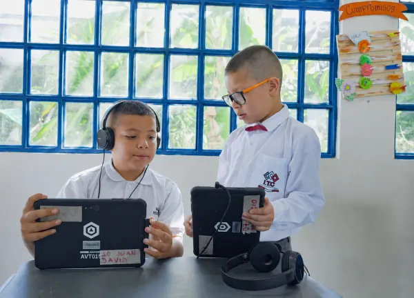 Two primary school students in uniform each work on their own AVACOM tablet, one of them wearing headphones, in a classroom with a glass block window