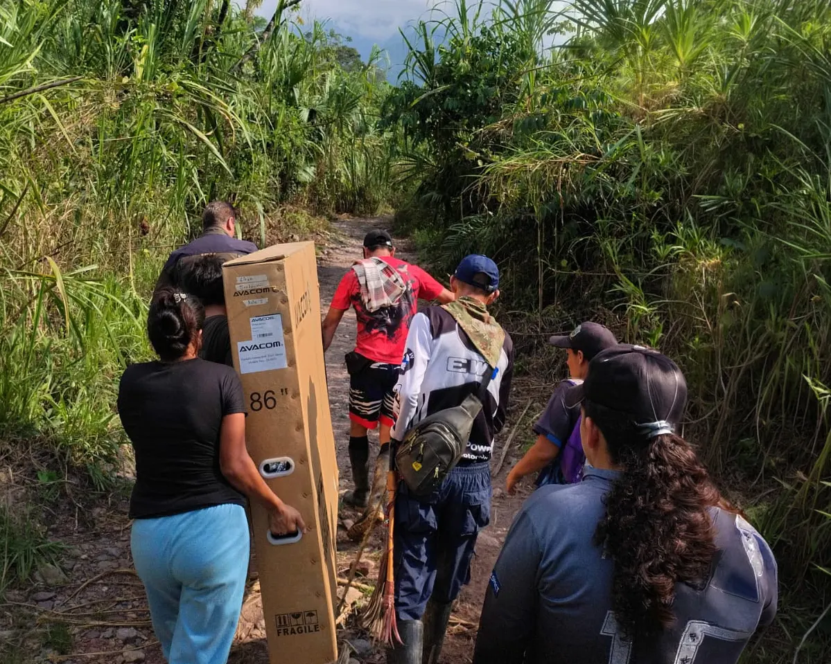 A group of people walking up a muddy track through dense tropical vegetation, one woman carrying an AVACOM box marked 86 inches by its handle
