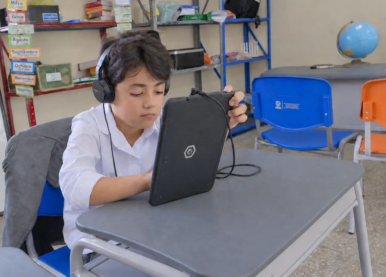 A student wearing headphones works intently on a tablet at a grey desk, with the months of the year on cards and a globe in the background