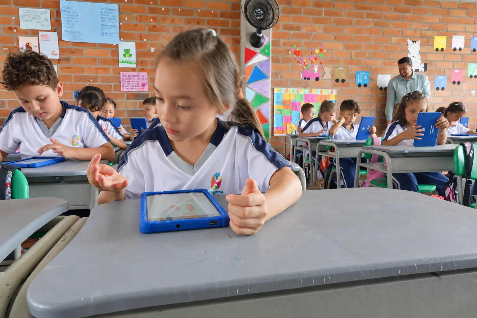 A student works with a tablet resting on her desk; behind her, hand-made posters on the brick wall and a teacher leaning over to help another student