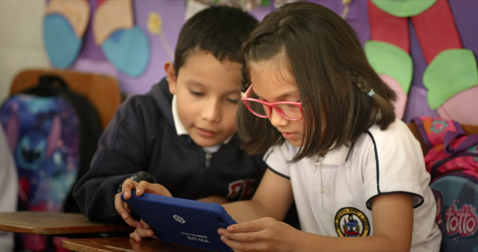 Two primary school students share an AVACOM tablet resting on their desk, with a colourful mural behind them