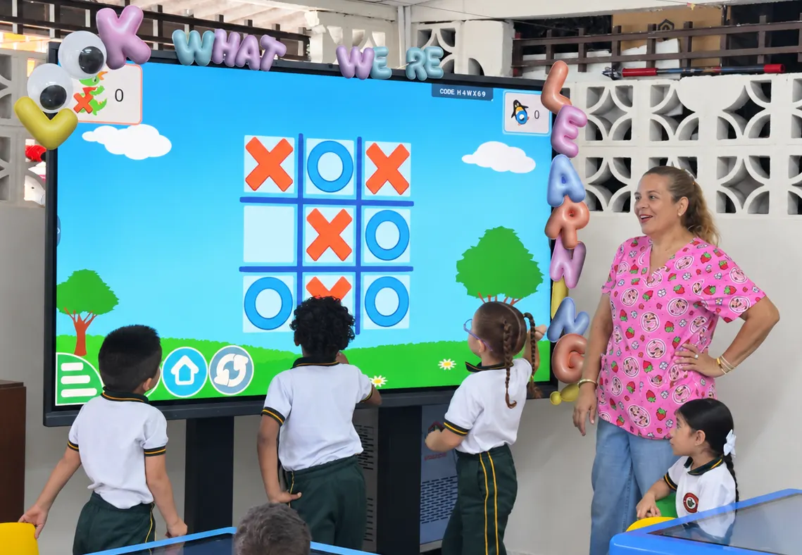 Three preschool children in front of an interactive screen showing a noughts-and-crosses game; one of them, wearing glasses, reaches out to the screen while the teacher looks on
