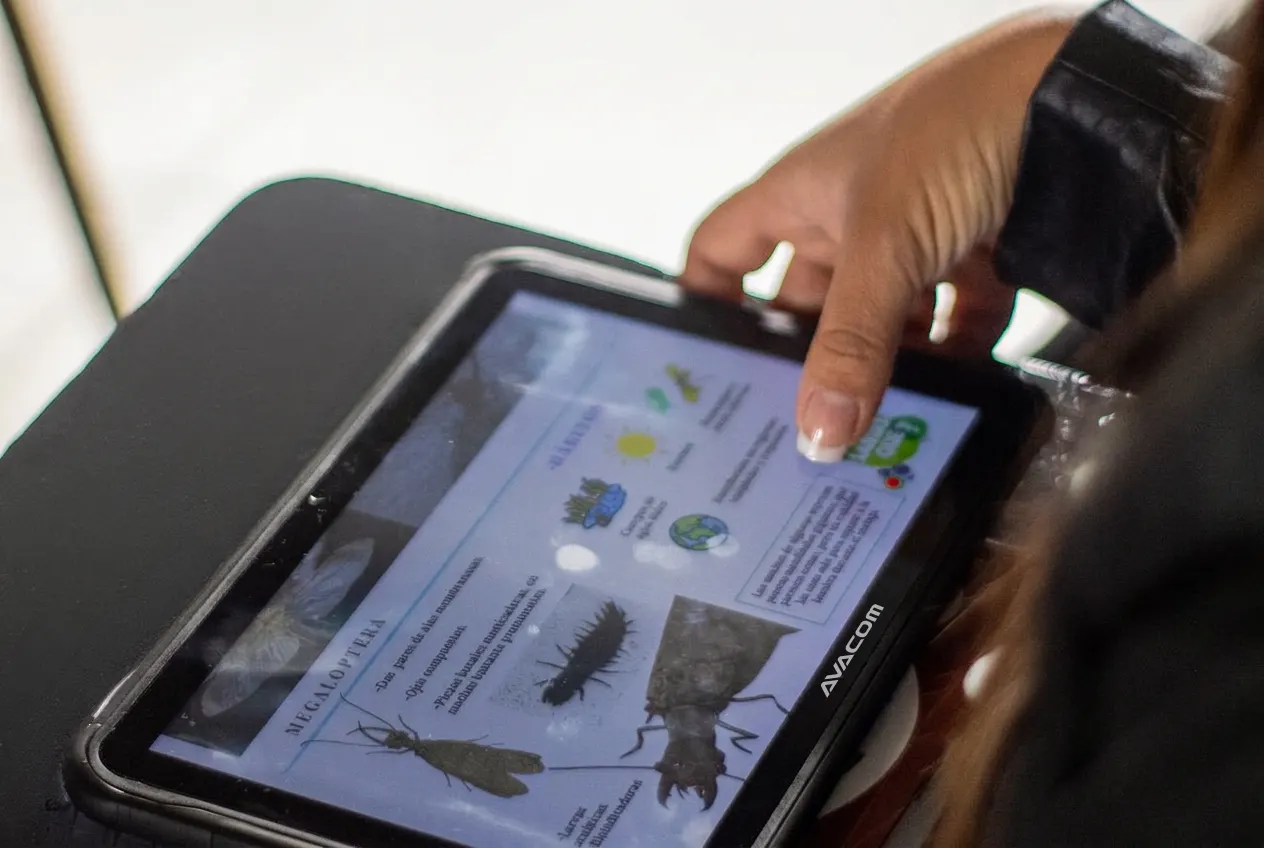 A woman's hand touching the screen of an AVACOM tablet in a black case, showing a natural sciences content card on the Megaloptera order